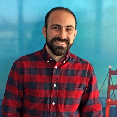 Portrait of Marinos Tsagkarakis smiling, wearing a red plaid shirt in front of a Golden Gate Bridge backdrop
